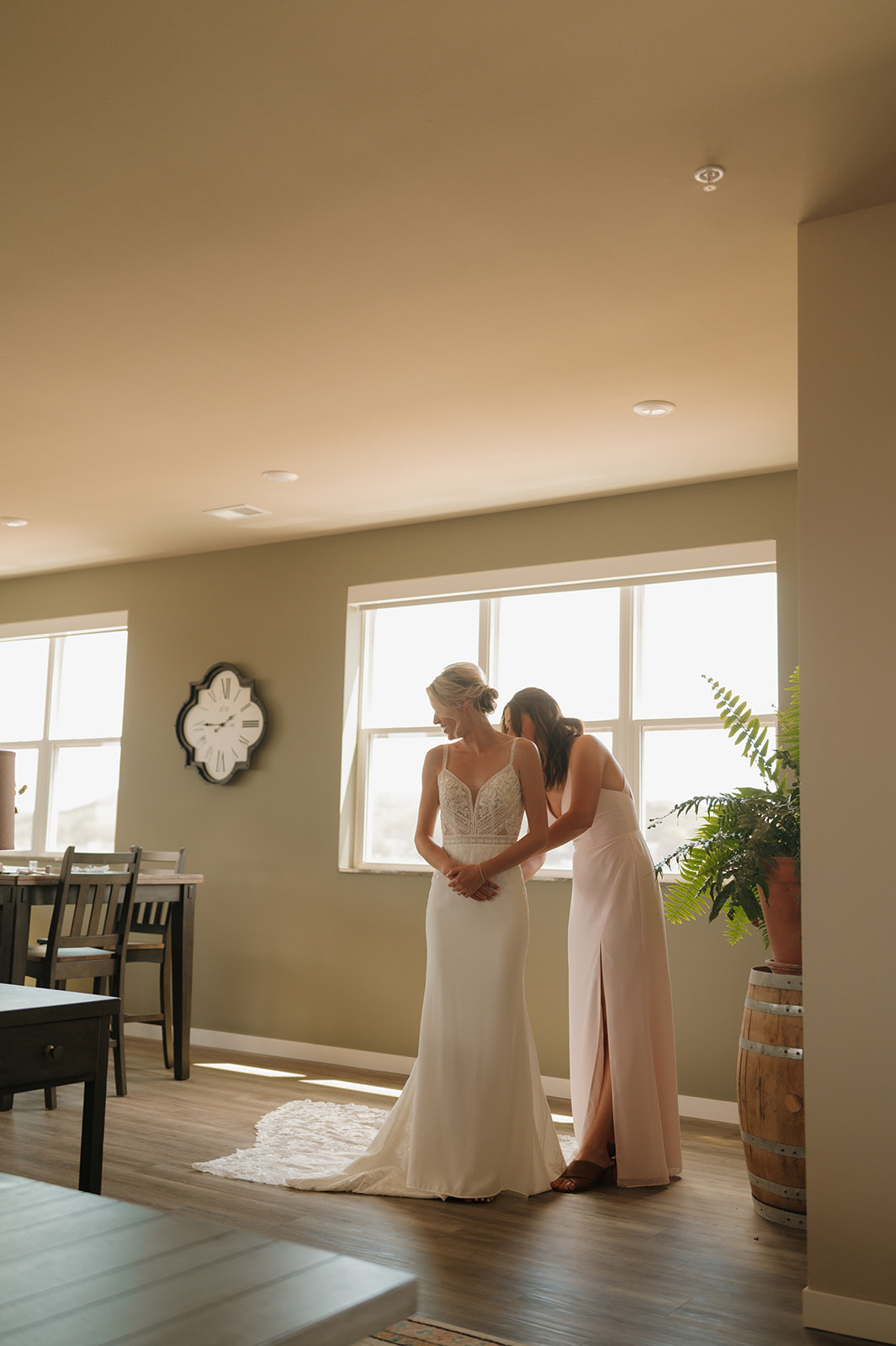 Bride smiling while getting buttoned into her gown with natural light flooding the room.