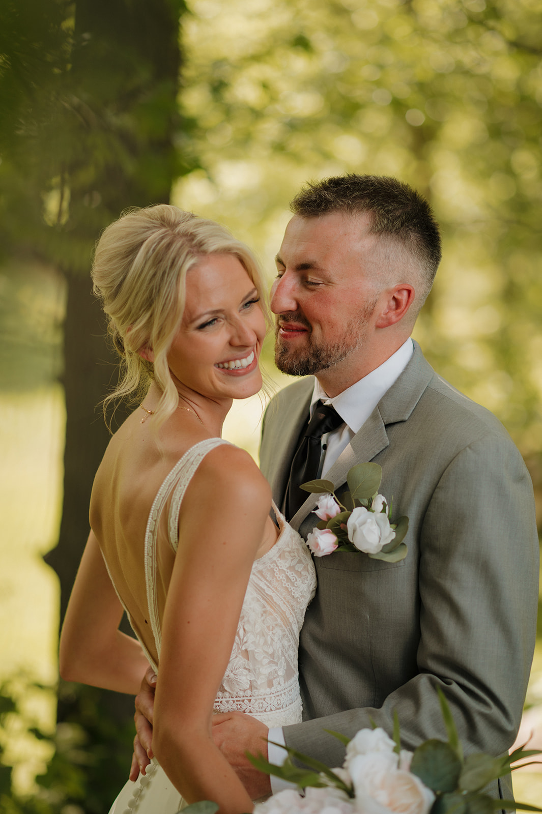 Groom lovingly gazes at the bride as they hold each other under leafy trees.