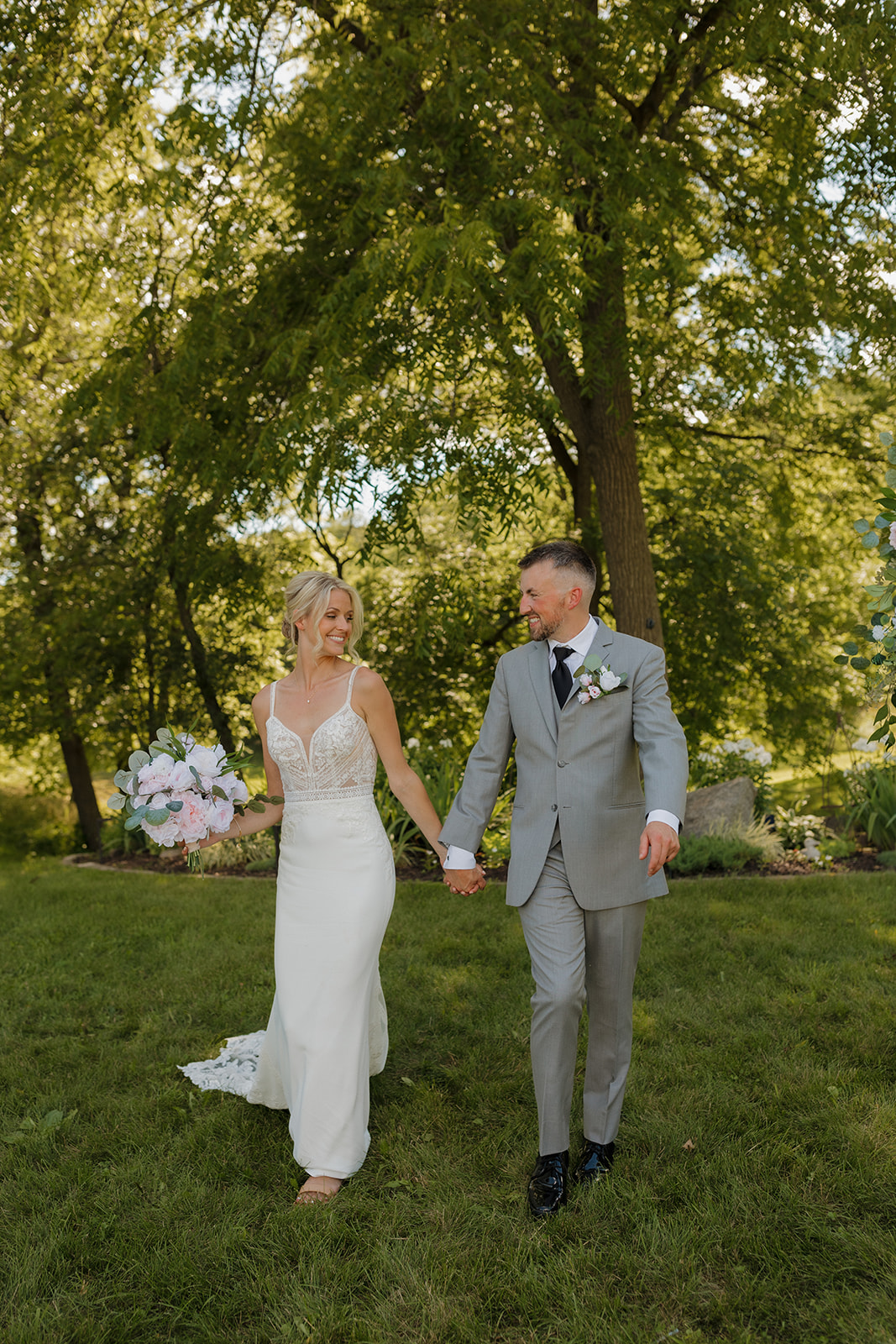 Bride and groom walk hand-in-hand through the grass, smiling at each other beneath tall trees.