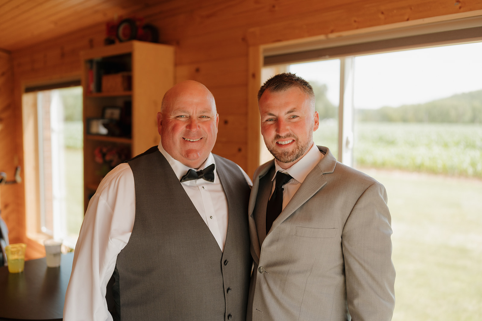 Groom poses with his father inside a cozy room with wood-paneled walls.