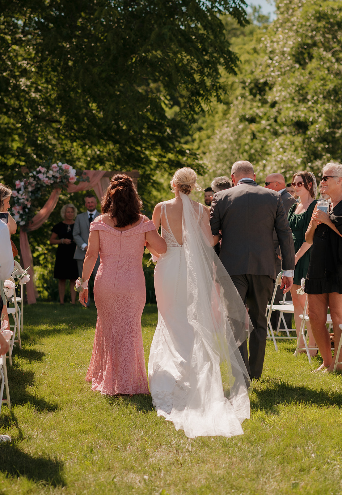 Bride walks arm in arm with her parents toward the altar for her outdoor vineyard wedding