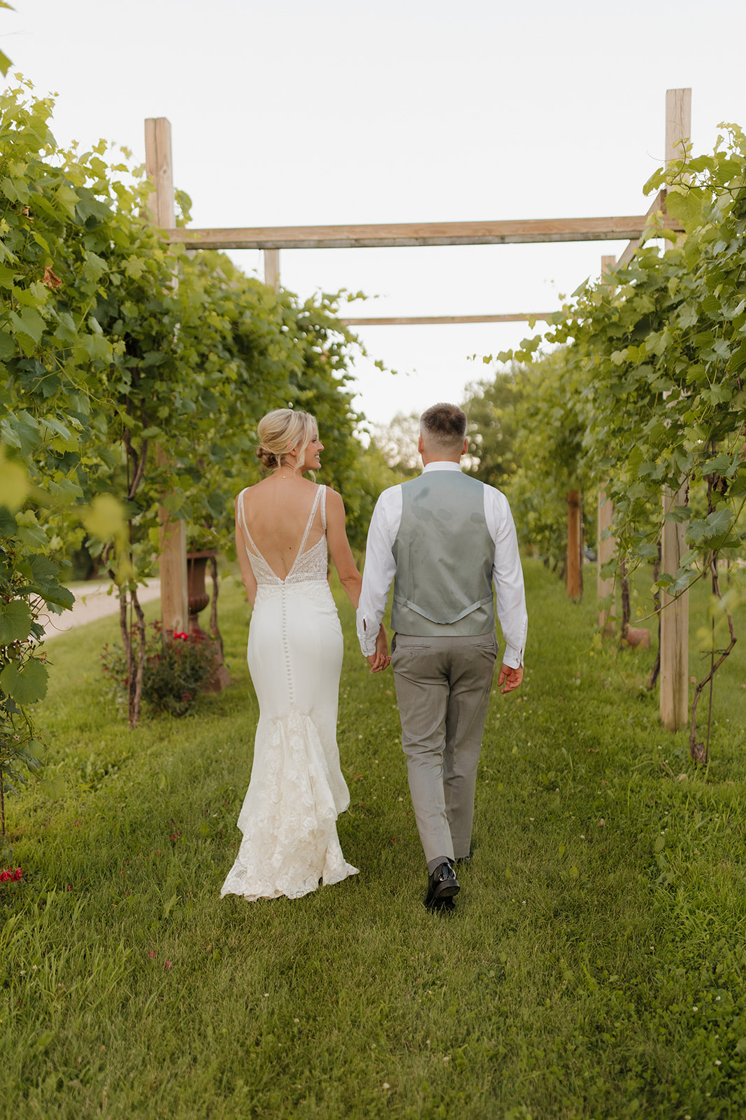 Bride and groom walk through lush vineyard rows under a wooden trellis at a Wisconsin winery wedding venue.