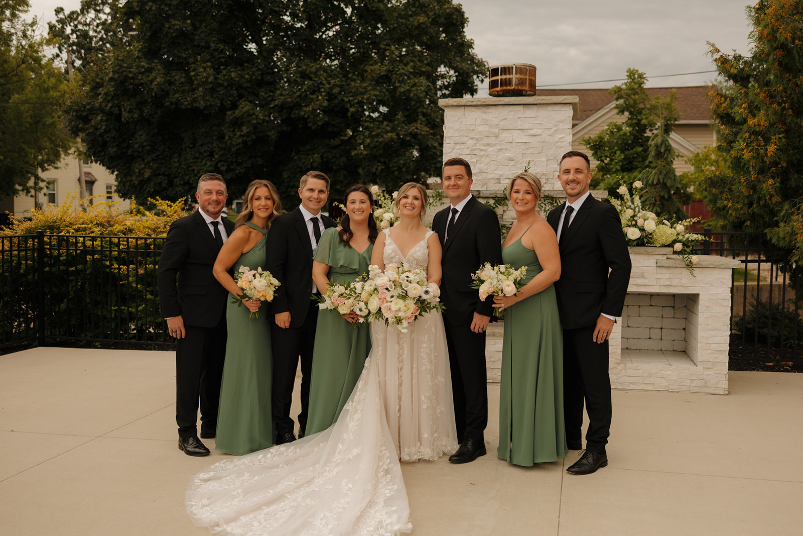 The bride and groom pose with their wedding party in front of a white fireplace altar, with green dresses and soft neutral florals adding elegant contrast—an image captured by wedding photographers in Wisconsin.