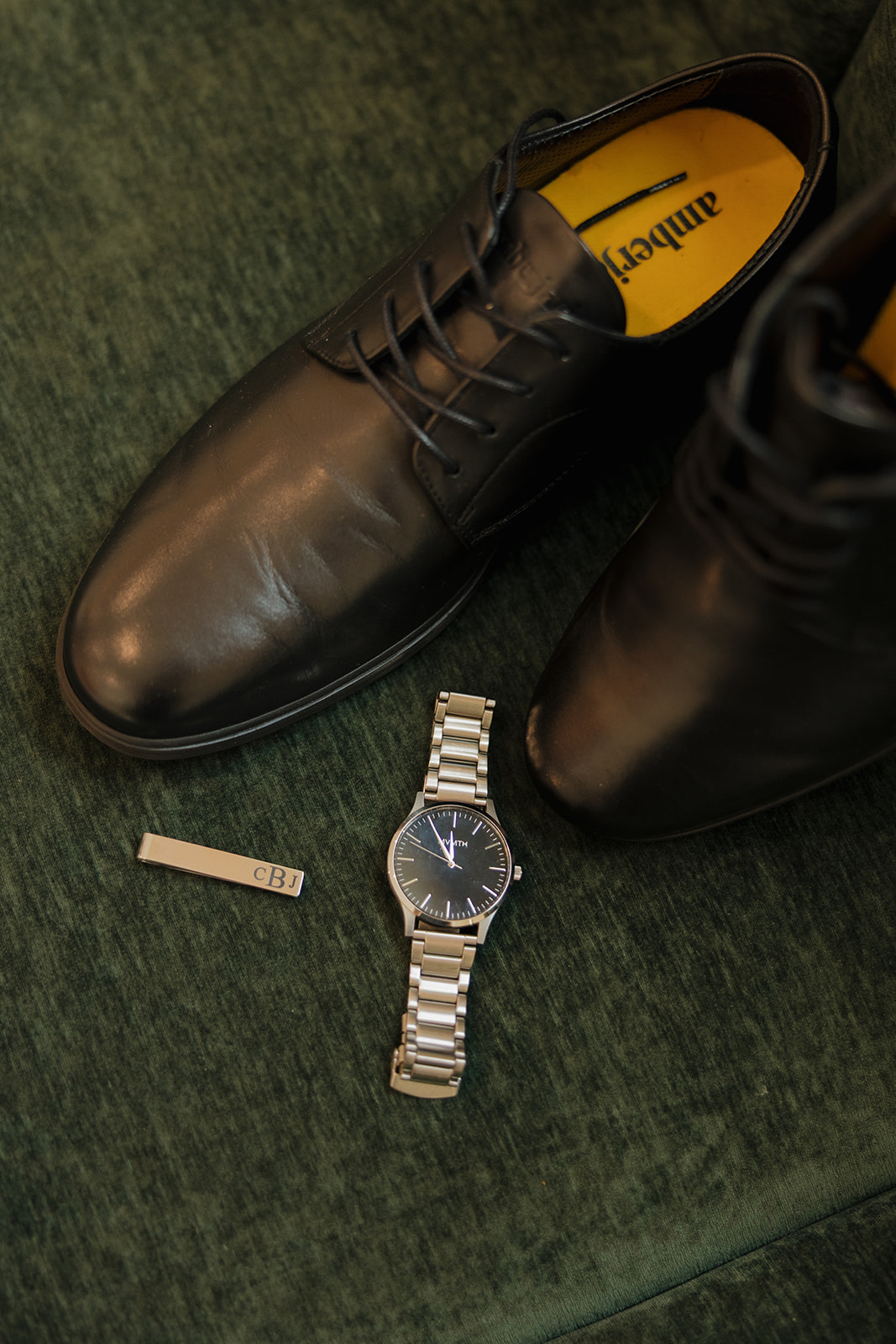 Classic black dress shoes, a silver watch, and personalized tie clip laid out on green velvet—groom details wedding photographers in Wisconsin love to capture.