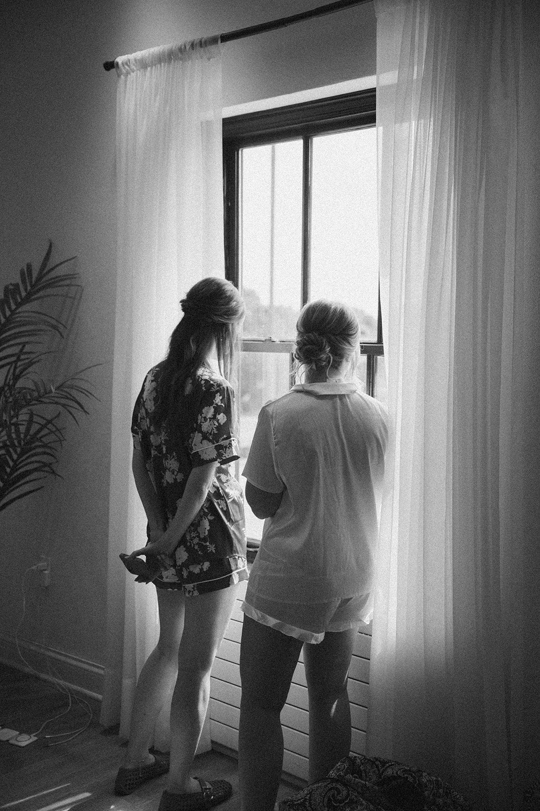 A candid black and white photo of two women standing by a window in matching pajama sets, soaking in the quiet before the wedding day begins.