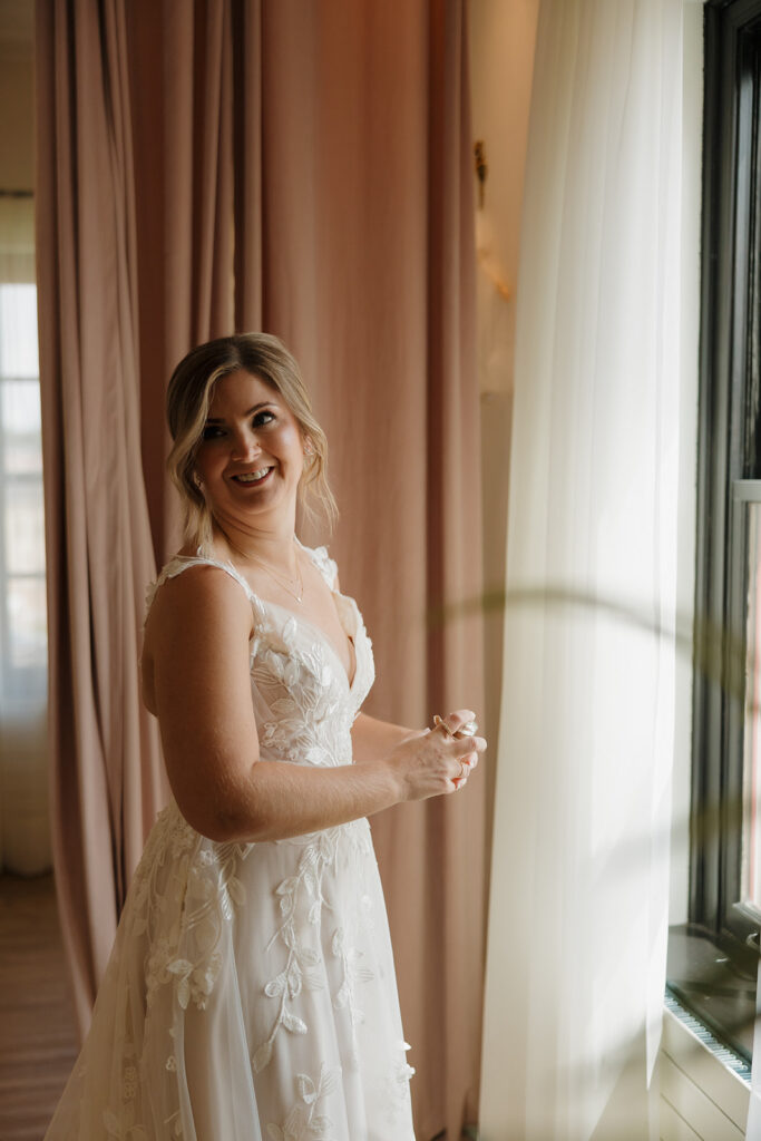 The bride stands near soft pink curtains and a window, smiling softly while clasping her necklace—a tender bridal moment captured by wedding photographers in Wisconsin.