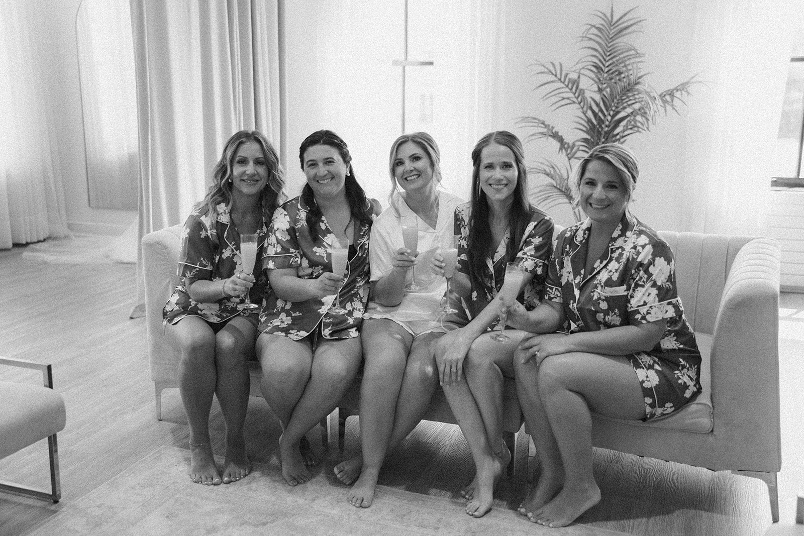 A cozy black and white portrait of the bride and bridesmaids in matching floral pajamas, seated barefoot on a plush couch and holding mimosas.