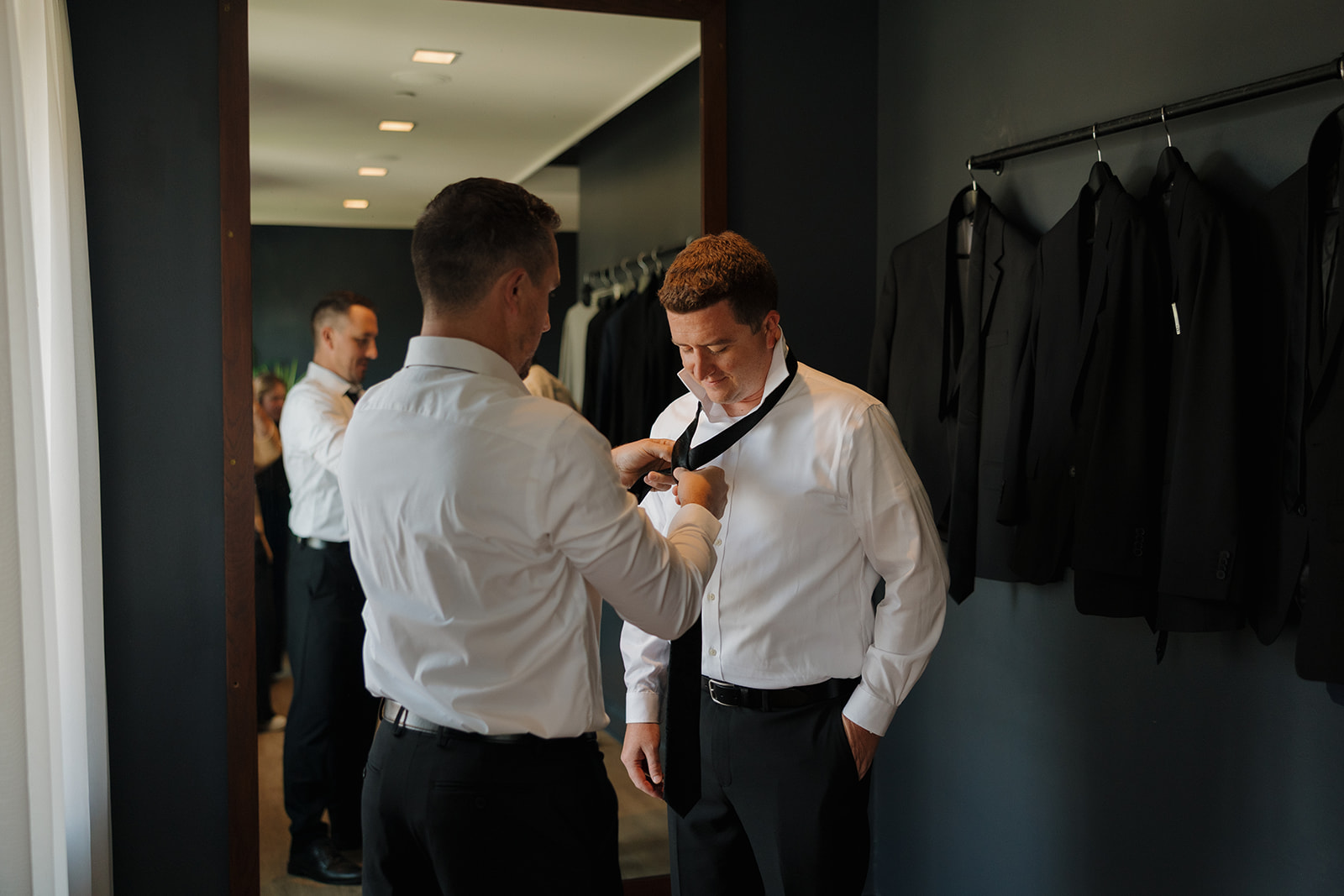 A groomsman helps the groom tie his black necktie in a sleek, modern prep room lined with black suits, a quiet moment captured by wedding photographers in Wisconsin.