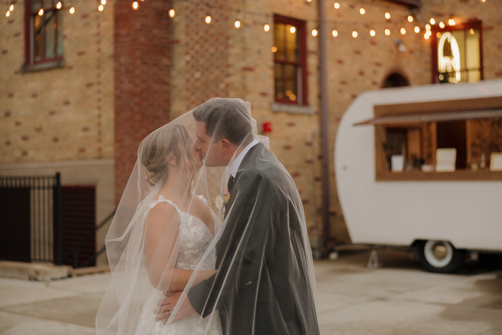 The bride and groom share a kiss beneath her veil in front of a vintage trailer bar, with string lights twinkling above—a dreamy scene captured by wedding photographers in Wisconsin.