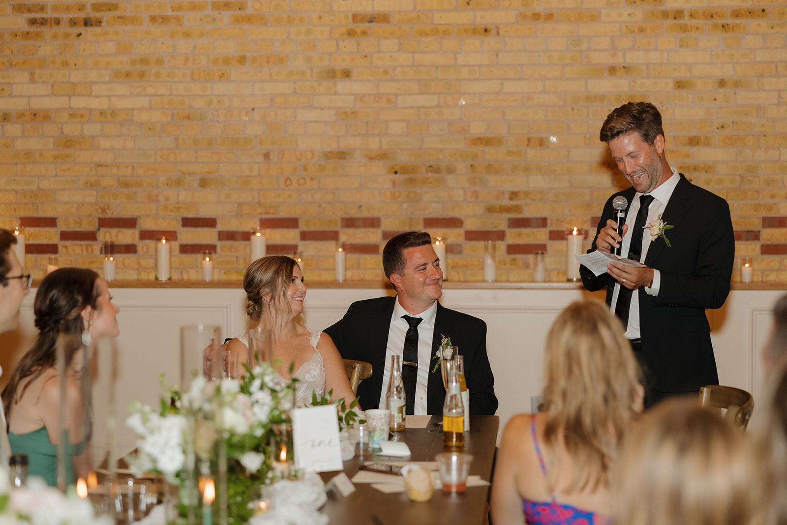 The groom laughs mid-toast while the bride beams beside him, surrounded by candles and soft brick warmth—a signature scene for wedding photographers in Wisconsin.