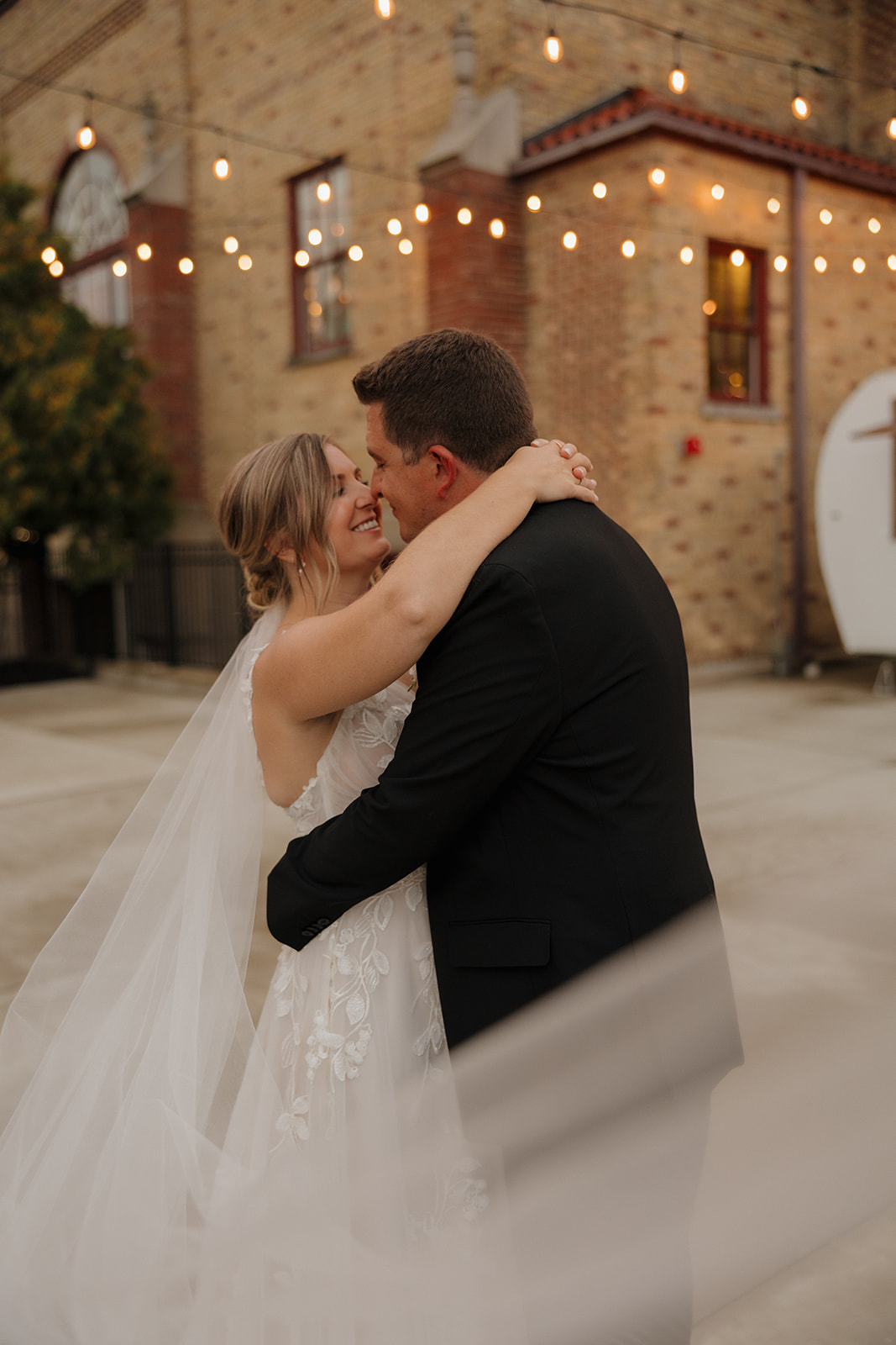 The bride and groom hold each other close beneath glowing string lights, wrapped in post-ceremony bliss.