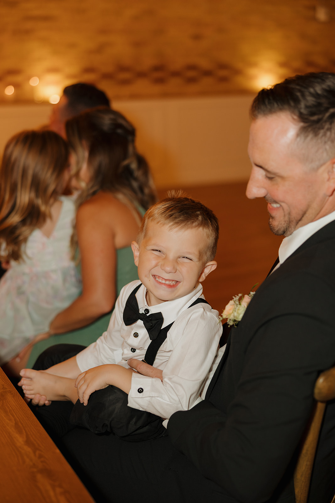 A little ring bearer grins ear-to-ear while sitting in a groomsman’s lap—tiny tux and all.