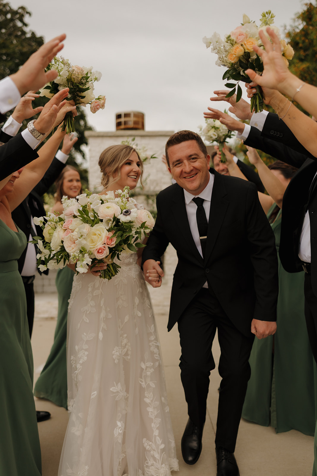 Newlyweds laugh and hold hands as they walk through a tunnel of bridesmaids and groomsmen raising bouquets, celebrating with pure joy and golden-hour glow.