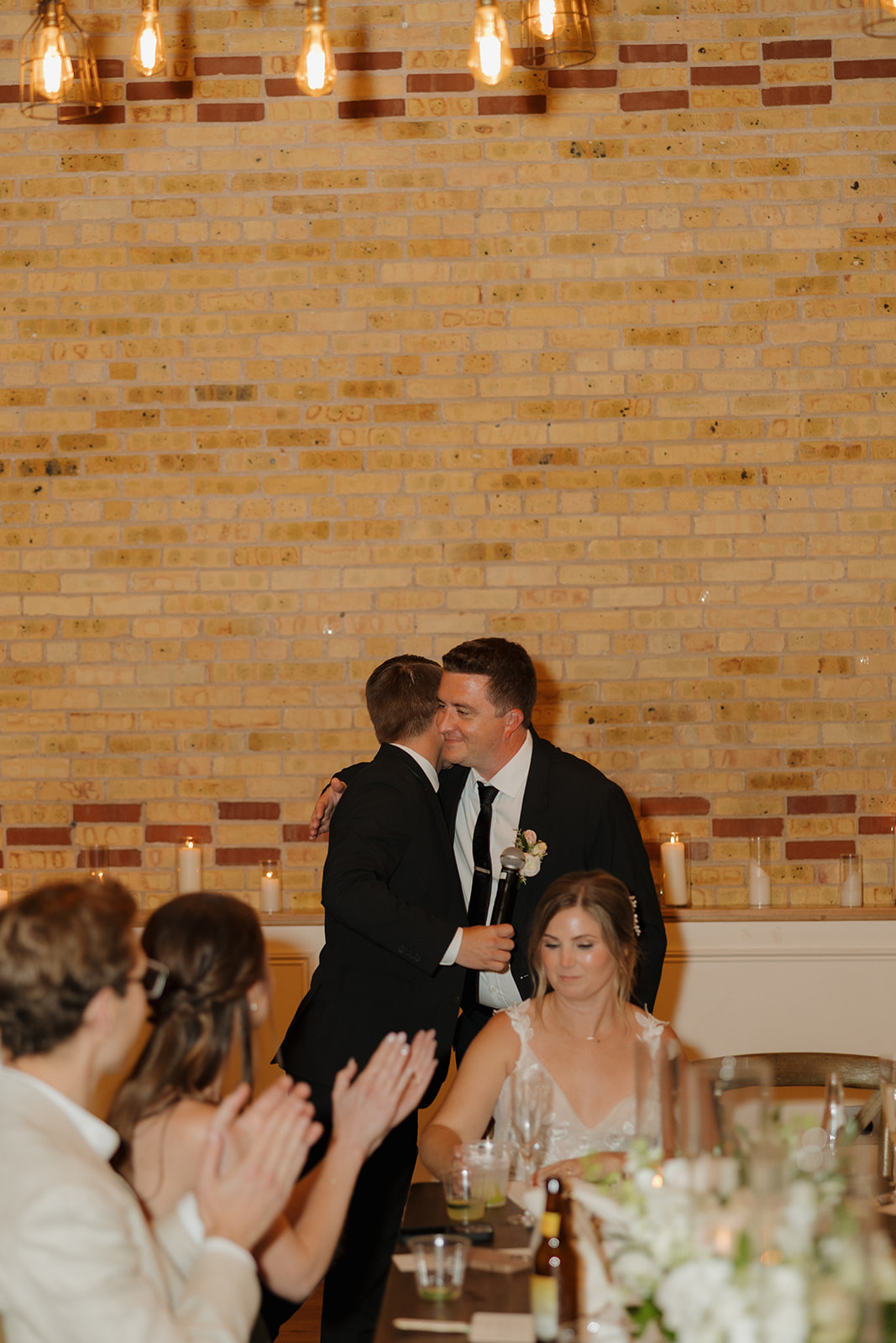 The groom embraces a guest after a touching speech, with guests applauding and smiles all around.