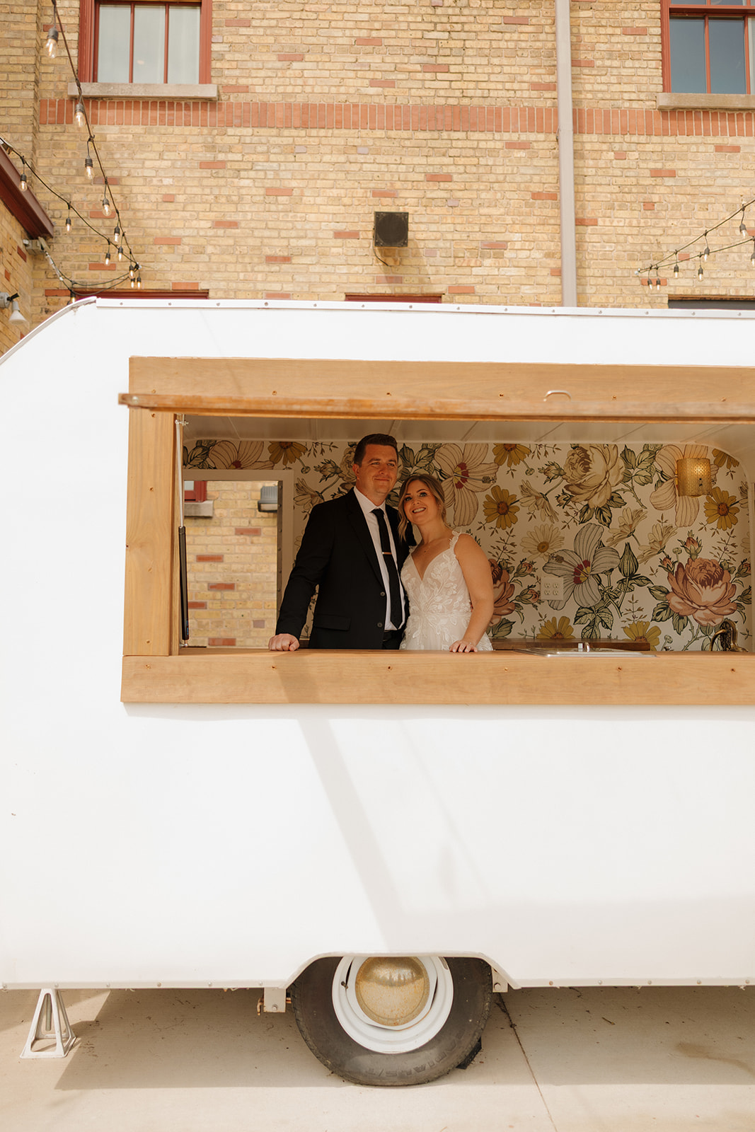 The newlyweds peek through a floral-wallpapered trailer window—quirky, charming, and so them.