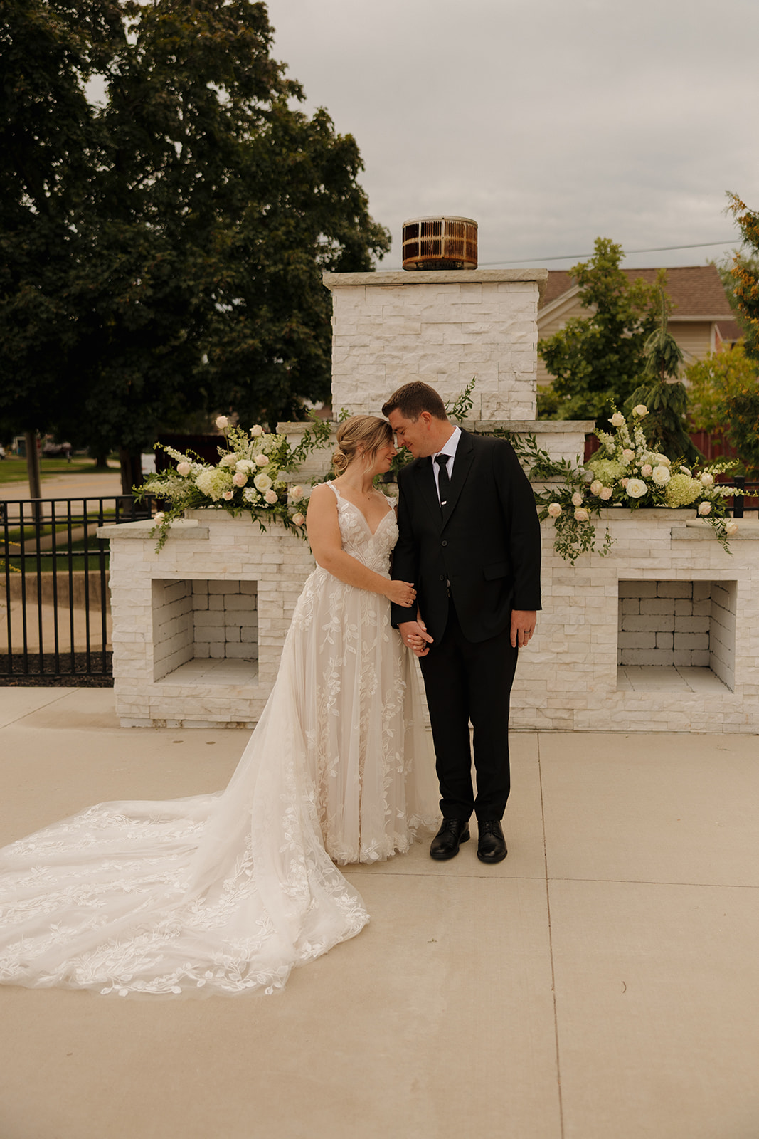 The bride and groom stand hand in hand in front of a white stone fireplace adorned with lush florals—timeless romance beautifully captured by wedding photographers in Wisconsin.