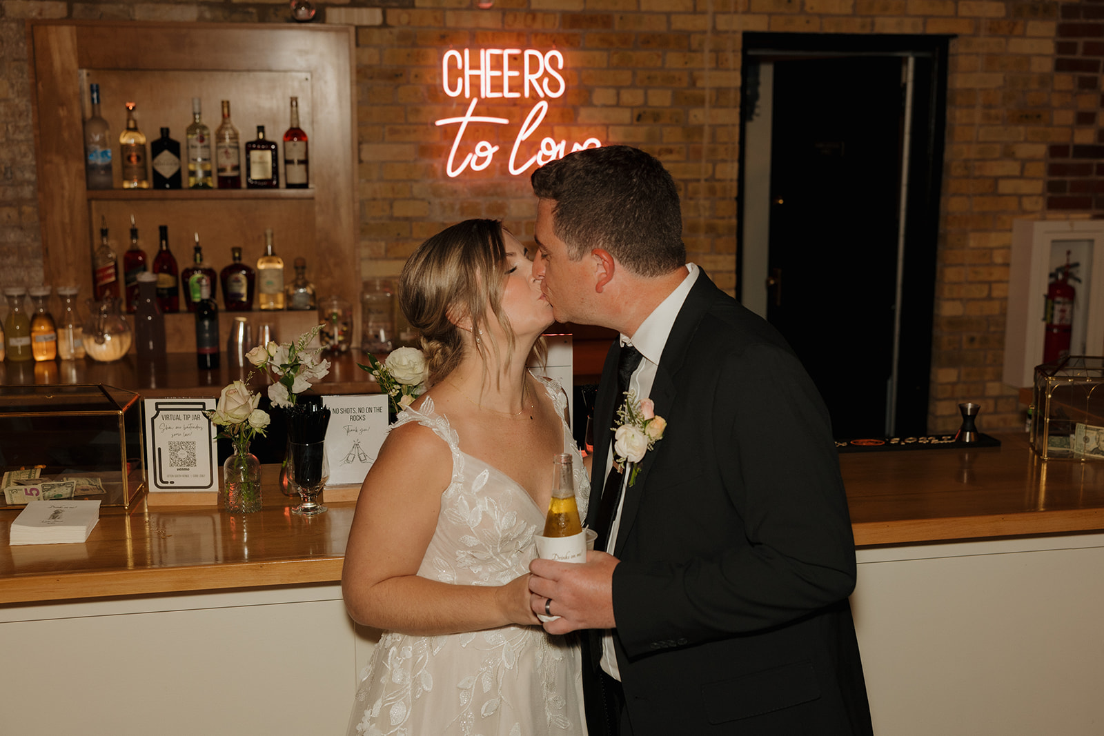 The couple shares a kiss in front of a bar with a neon “Cheers to Love” sign, toasting to forever with drinks in hand.