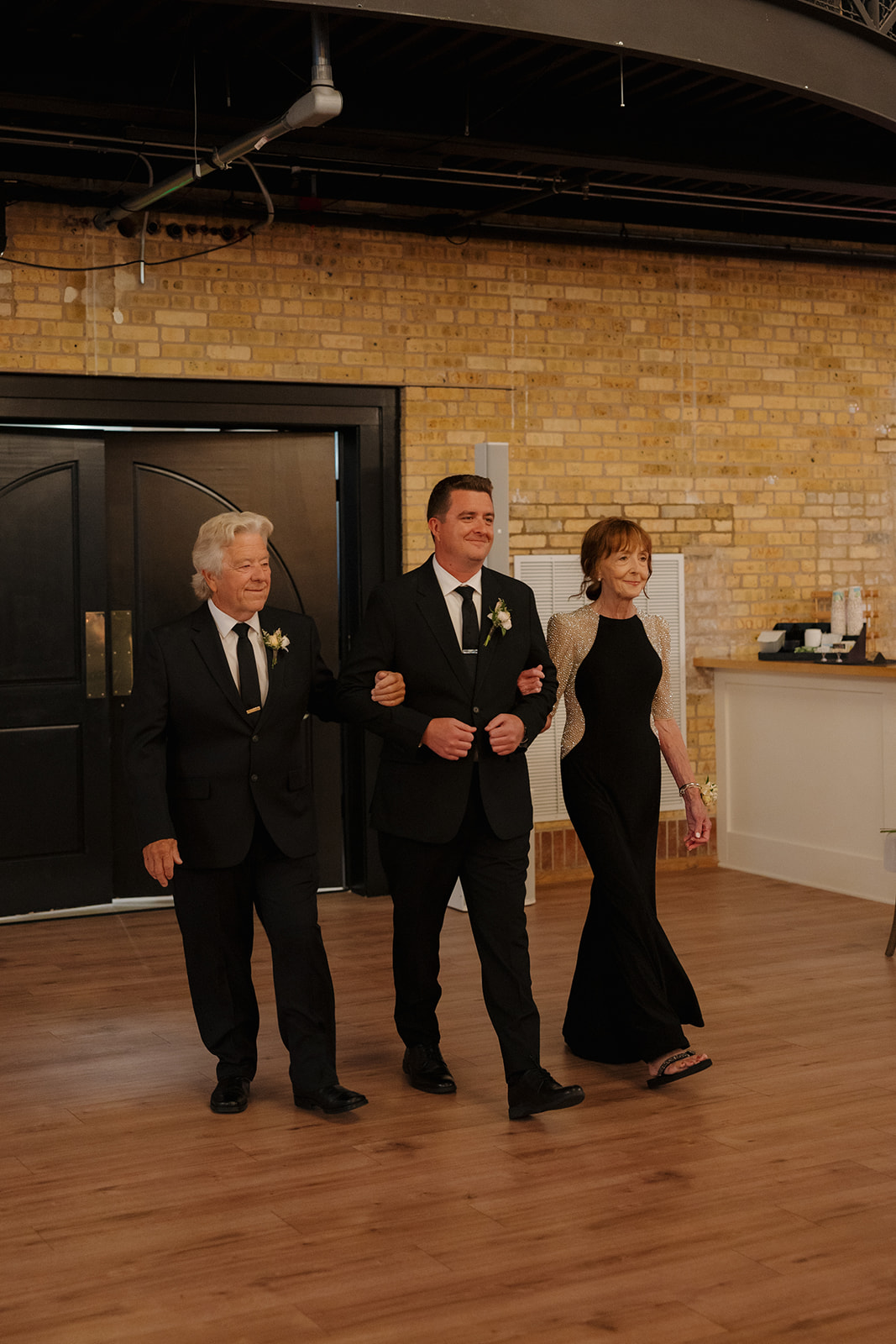 The groom walks arm-in-arm with both parents, smiling as he enters the reception space lined with candles and exposed brick walls.
