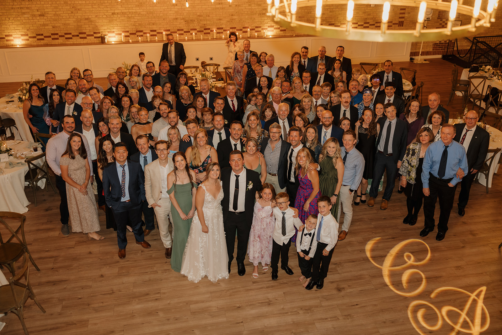 A big group photo of every guest, all smiling around the couple beneath glowing chandeliers and a monogrammed dance floor.