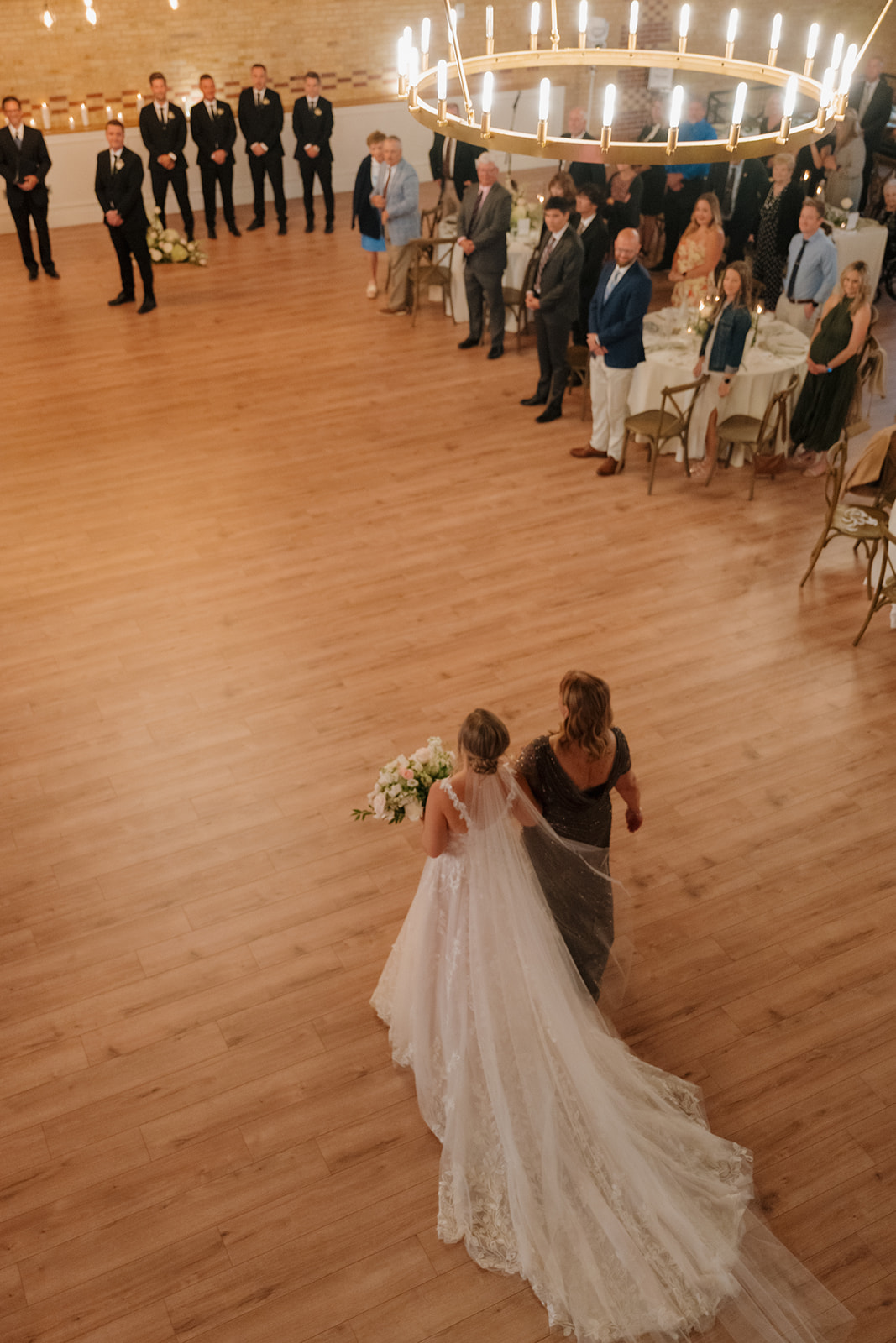 The bride makes her grand entrance, walking down the aisle with her mother as guests look on—an emotional moment full of anticipation.