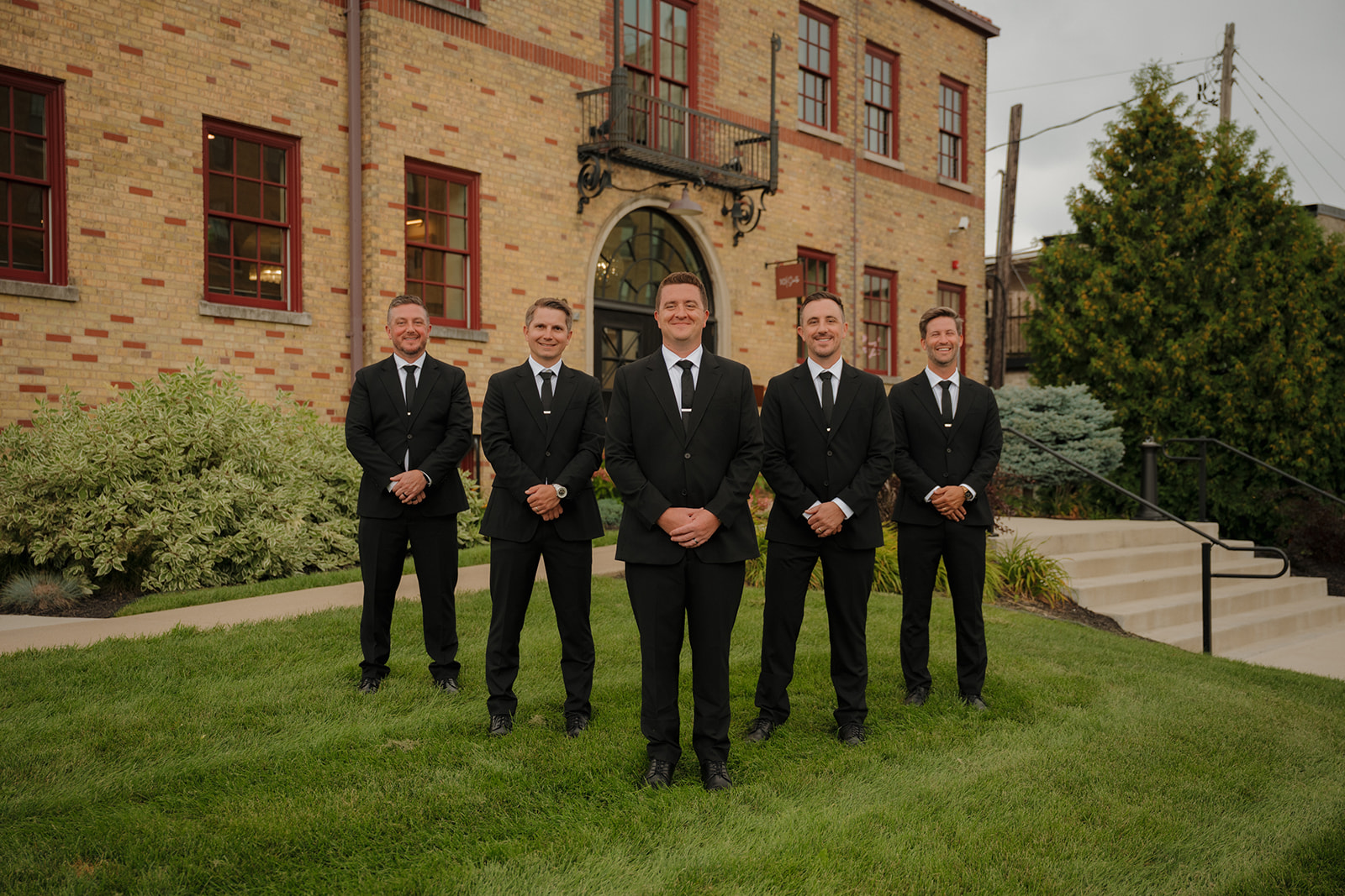 A classic group portrait of the groom and his groomsmen standing in front of a vintage brick venue, suited up and ready for the day.