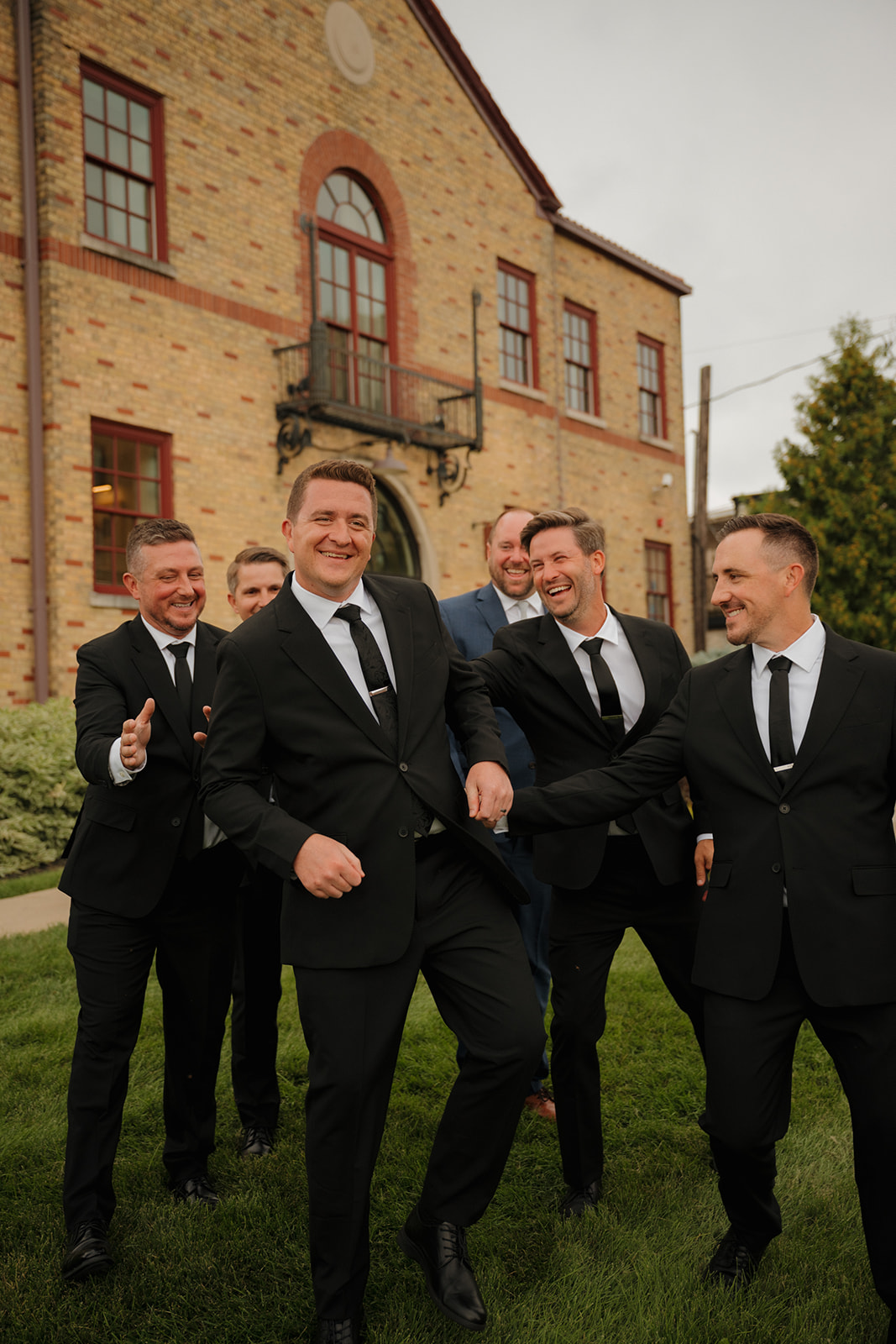 The groom shares a laugh with his groomsmen in front of the brick venue, a playful and candid moment that shows off the bond behind the suits.