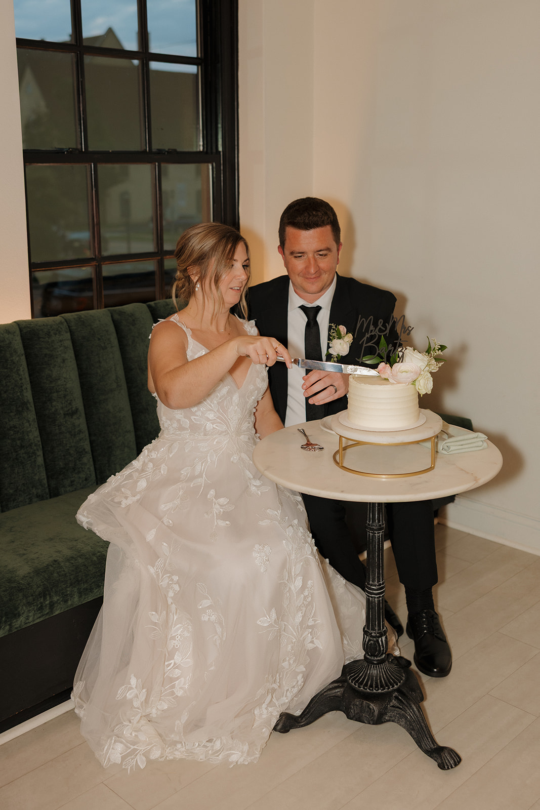Seated side-by-side on a plush green bench, the newlyweds cut their wedding cake—sweet, simple, and full of charm.
