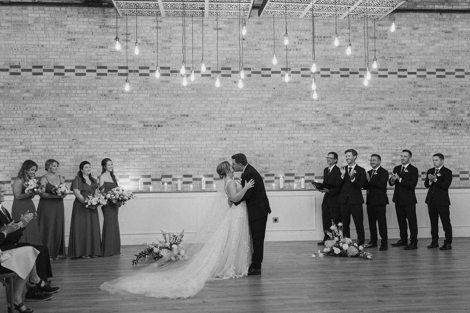 The bride and groom share their first kiss as a married couple under a canopy of hanging lights, surrounded by cheering friends and family.