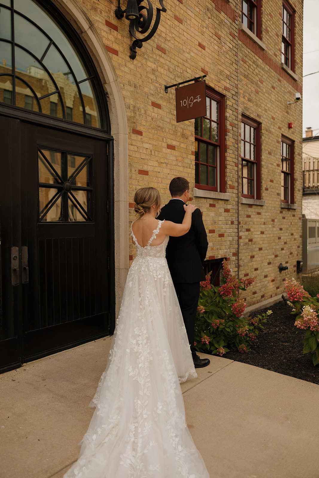 The bride approaches the groom from behind for their first look outside a historic brick venue, her lace train trailing behind her.