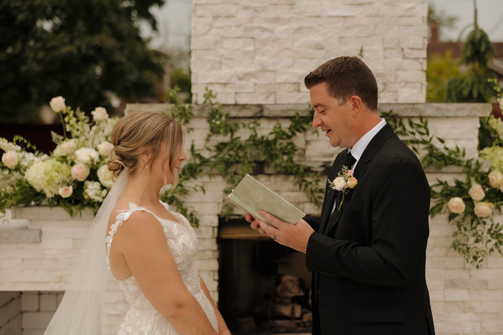 The groom reads heartfelt vows to the bride in front of a floral-adorned white fireplace, with a soft smile and green vow book in hand.
