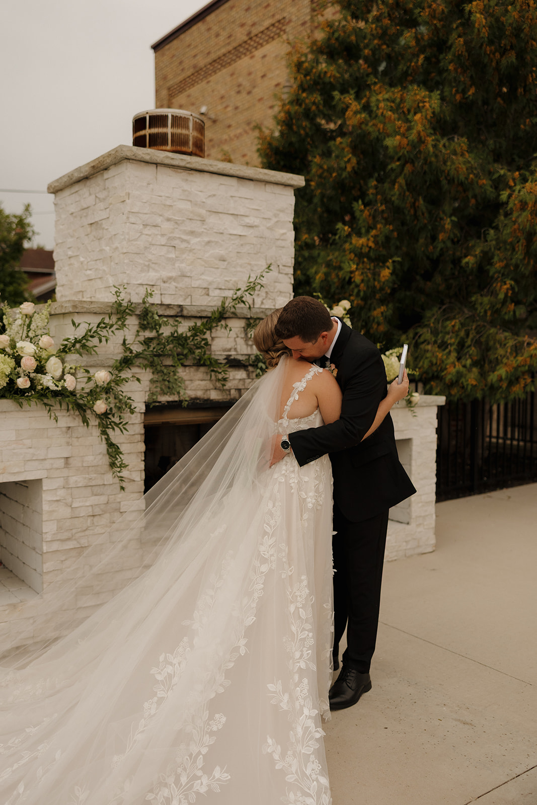 The bride and groom share a tearful embrace after exchanging vows in front of a white fireplace, surrounded by fresh greenery and soft blooms.