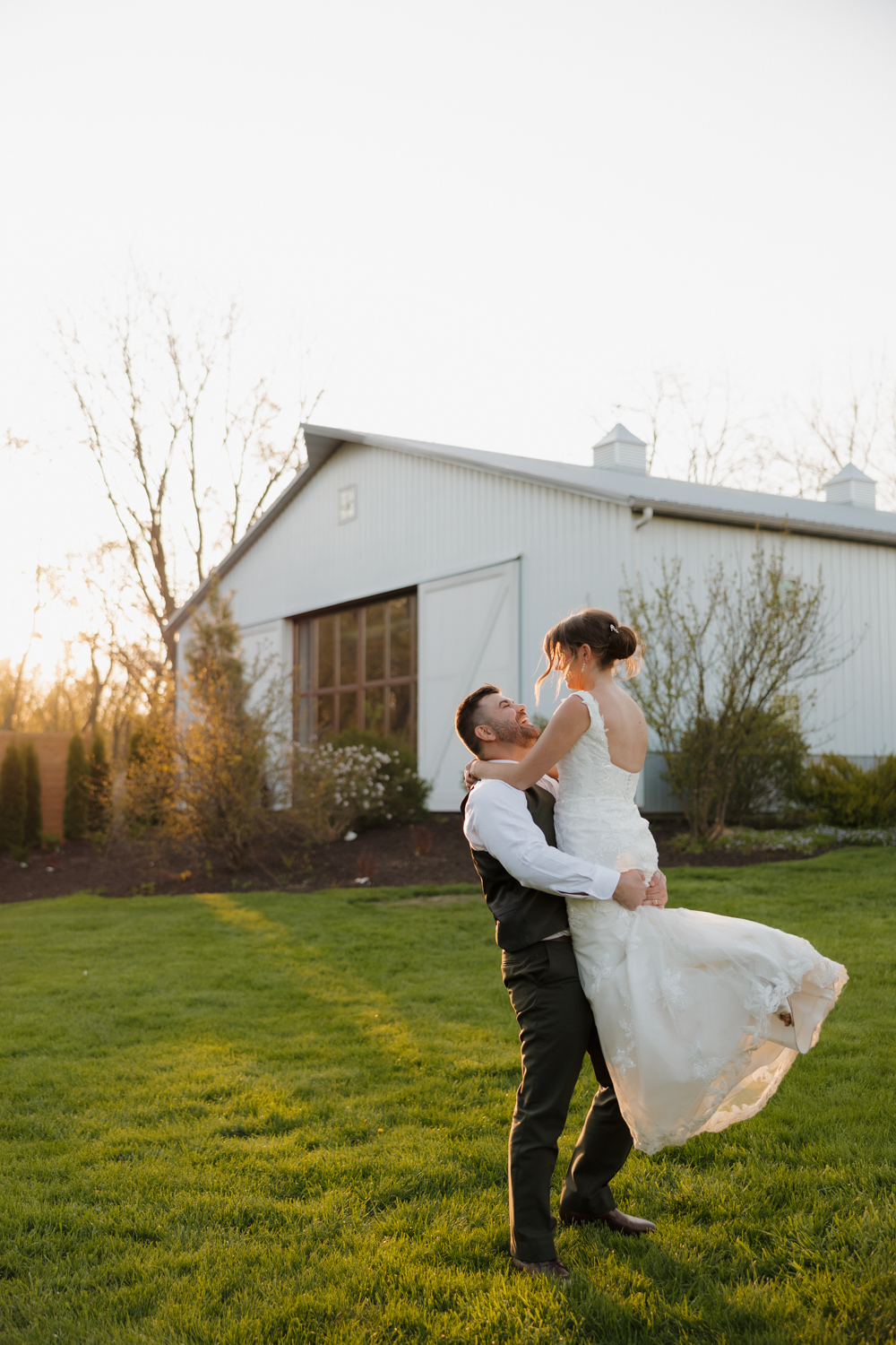 Groom lifting the bride during golden hour outside Boxed and Burlap Delavan with the white barn glowing in the background.
