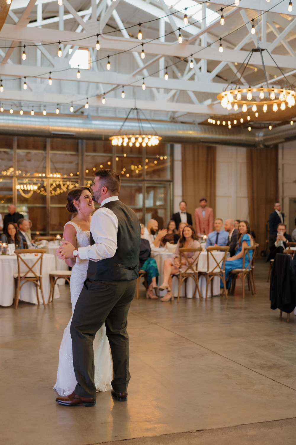 Bride and groom sharing their first dance inside Boxed and Burlap Delavan beneath chandeliers and exposed white beams.
