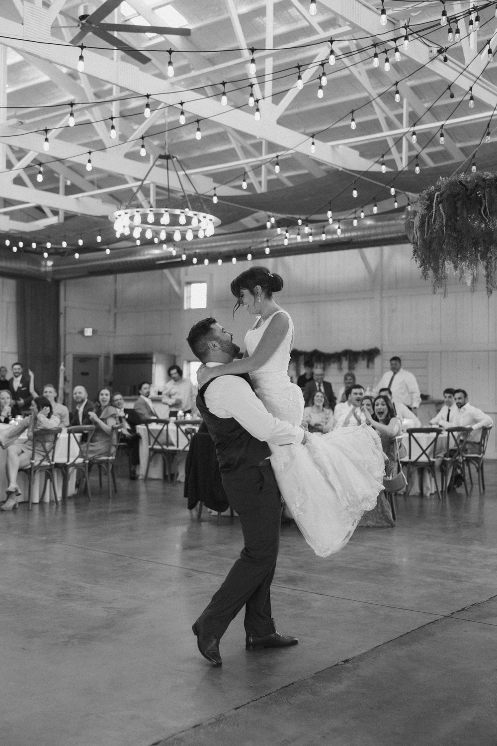 Black and white photo of groom lifting the bride on the dance floor at Boxed and Burlap Delavan as guests cheer beneath string lights.