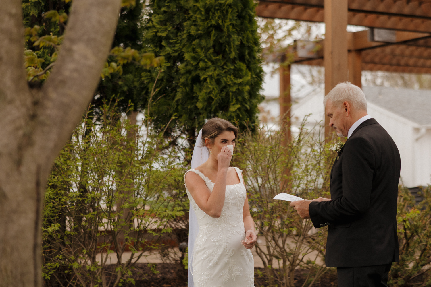 Bride wiping away tears while her father reads during an emotional outdoor ceremony.
