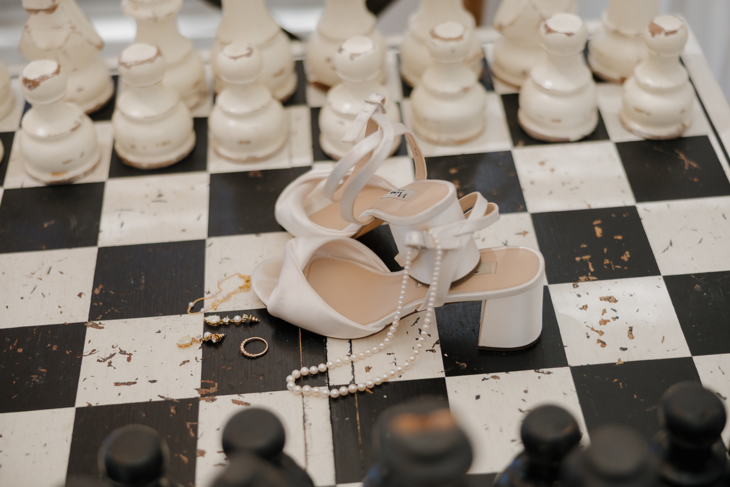 White bridal heels, pearl necklace, and gold jewelry styled on a black and white checkered table at Boxed and Burlap Delavan.