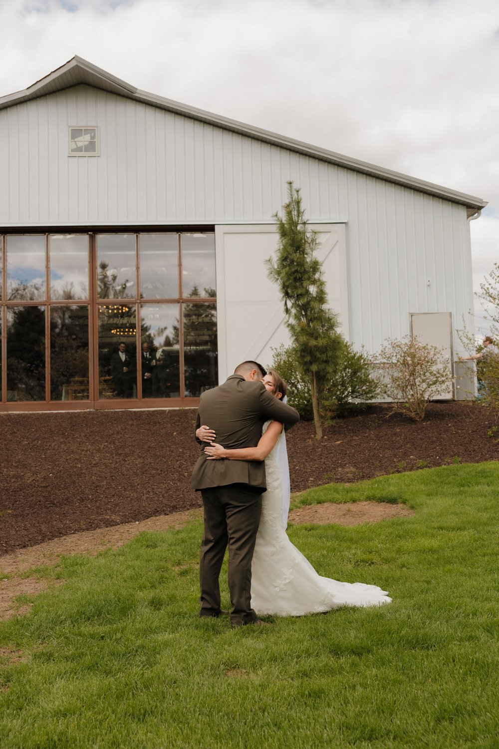 Bride and groom sharing a quiet embrace outside Boxed and Burlap Delavan with the white barn and large windows behind them.