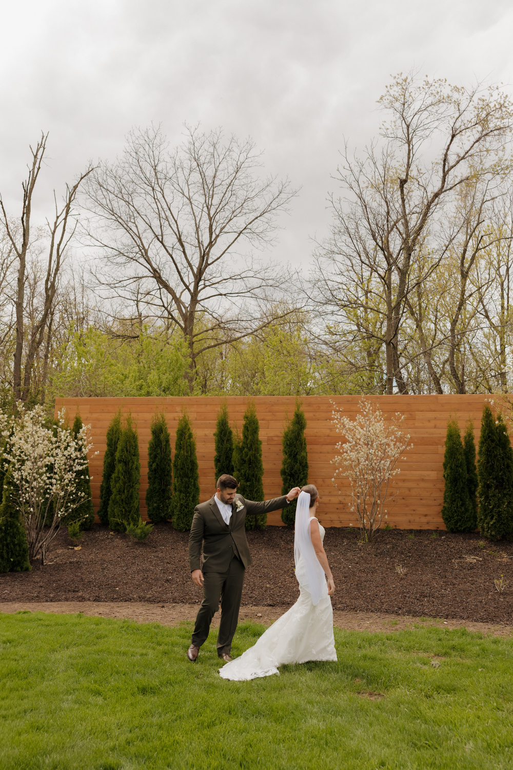 Bride and groom walking hand in hand outside Boxed and Burlap Delavan with a wooden privacy wall and manicured greenery in the background.