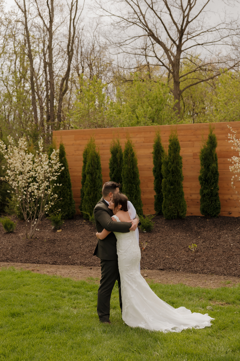 Bride and groom embracing on the lawn in front of a modern wooden fence and landscaped greenery.