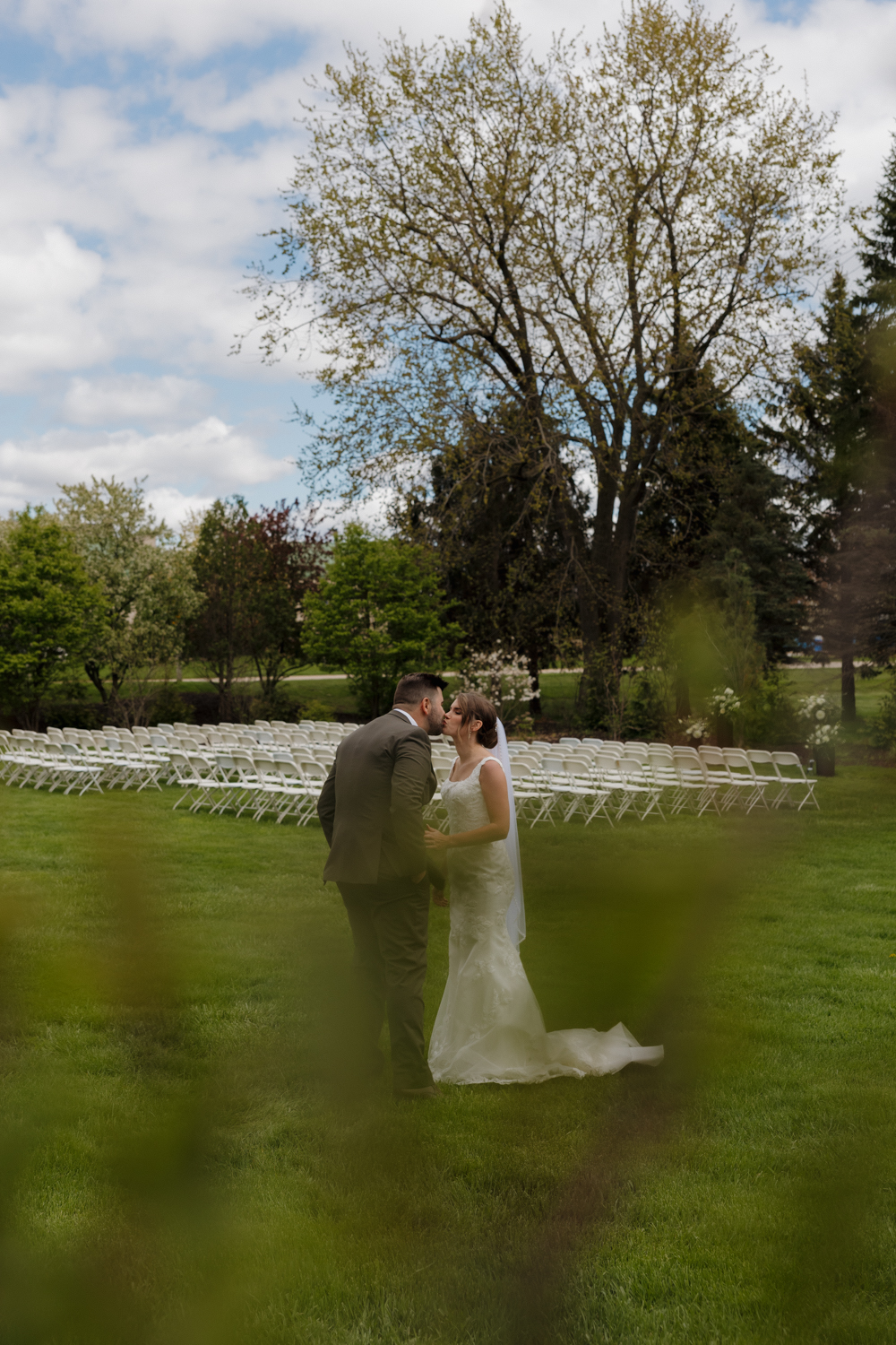 Bride and groom share a kiss on the lawn after their outdoor ceremony at Boxed and Burlap Delavan, surrounded by rows of white chairs and spring greenery.