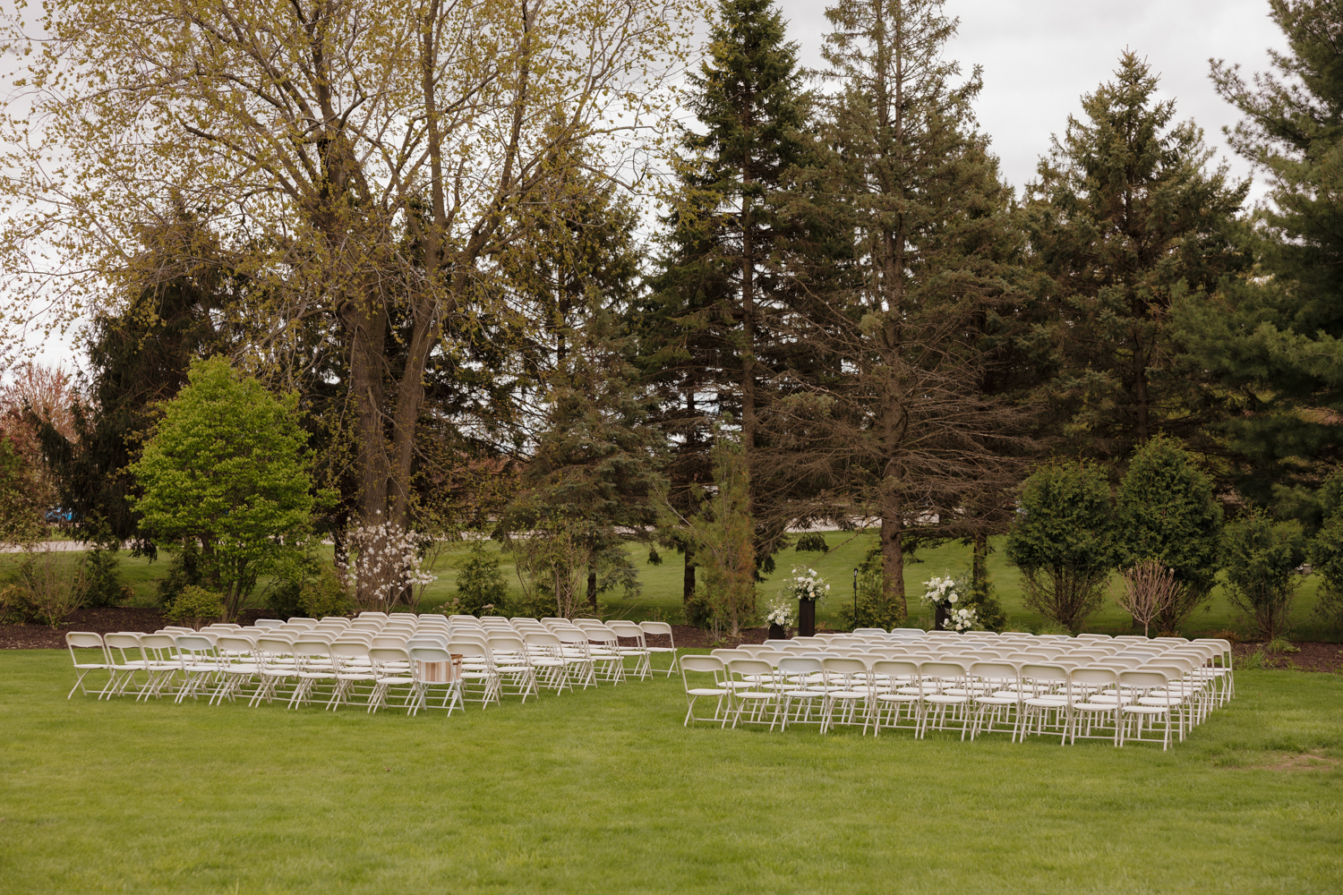 Outdoor ceremony setup at Boxed and Burlap Delavan with rows of white chairs arranged on the lawn beneath tall trees.