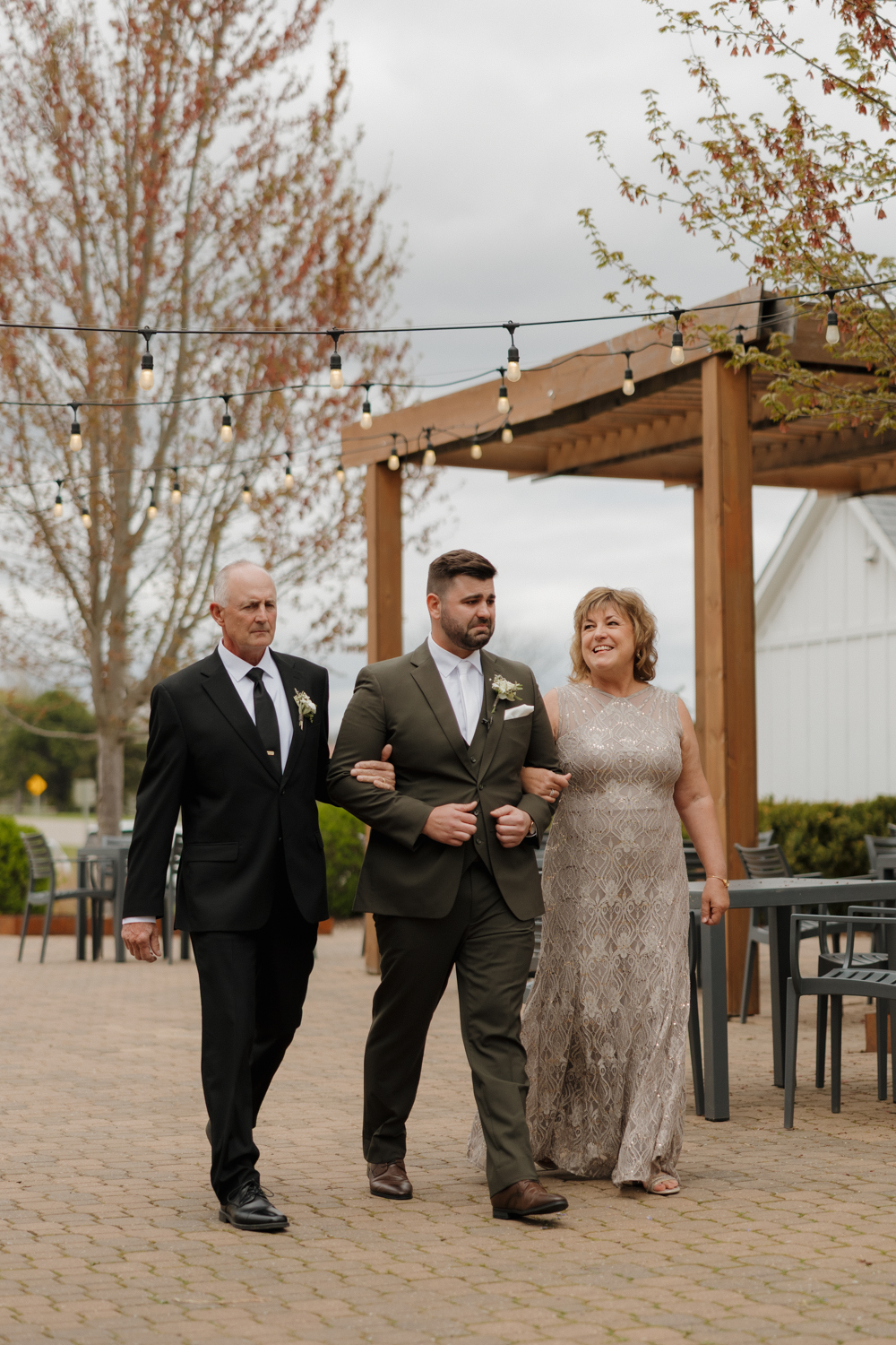 Groom walking arm in arm with his parents beneath café string lights outside Boxed and Burlap Delavan.