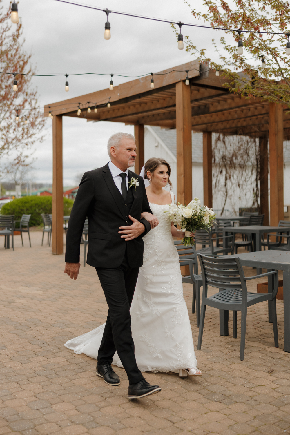 Bride walking arm in arm with her father outside Boxed and Burlap Delavan beneath café string lights and a wooden pergola.