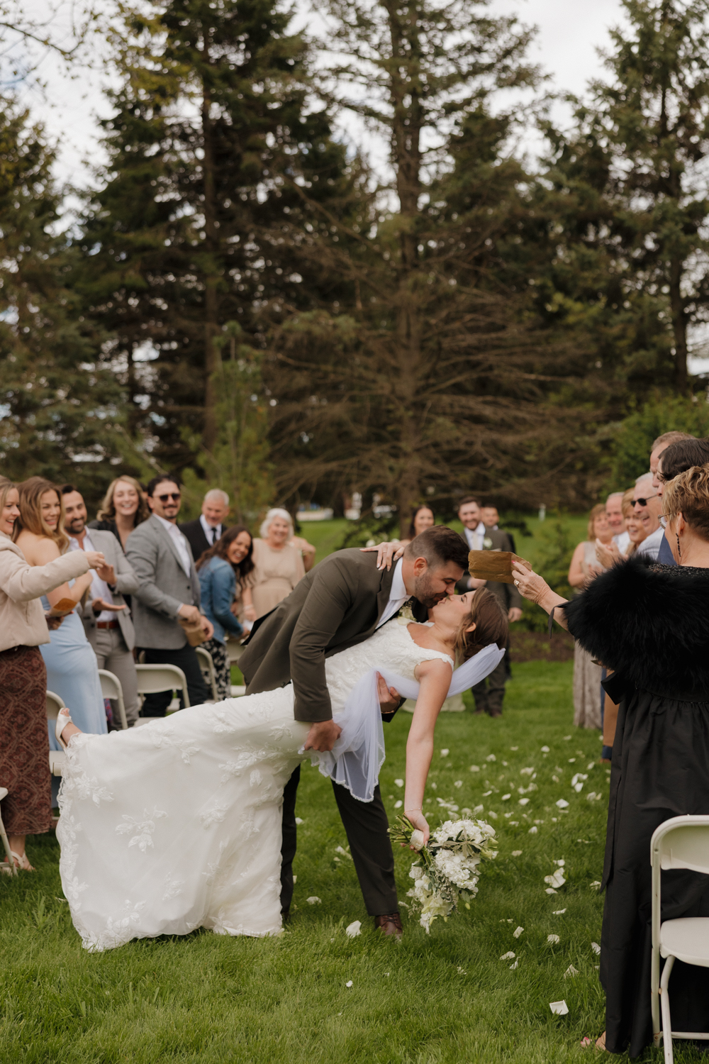 Bride and groom sharing a celebratory dip and kiss down the outdoor aisle as guests toss flower petals at Boxed and Burlap Delavan.
