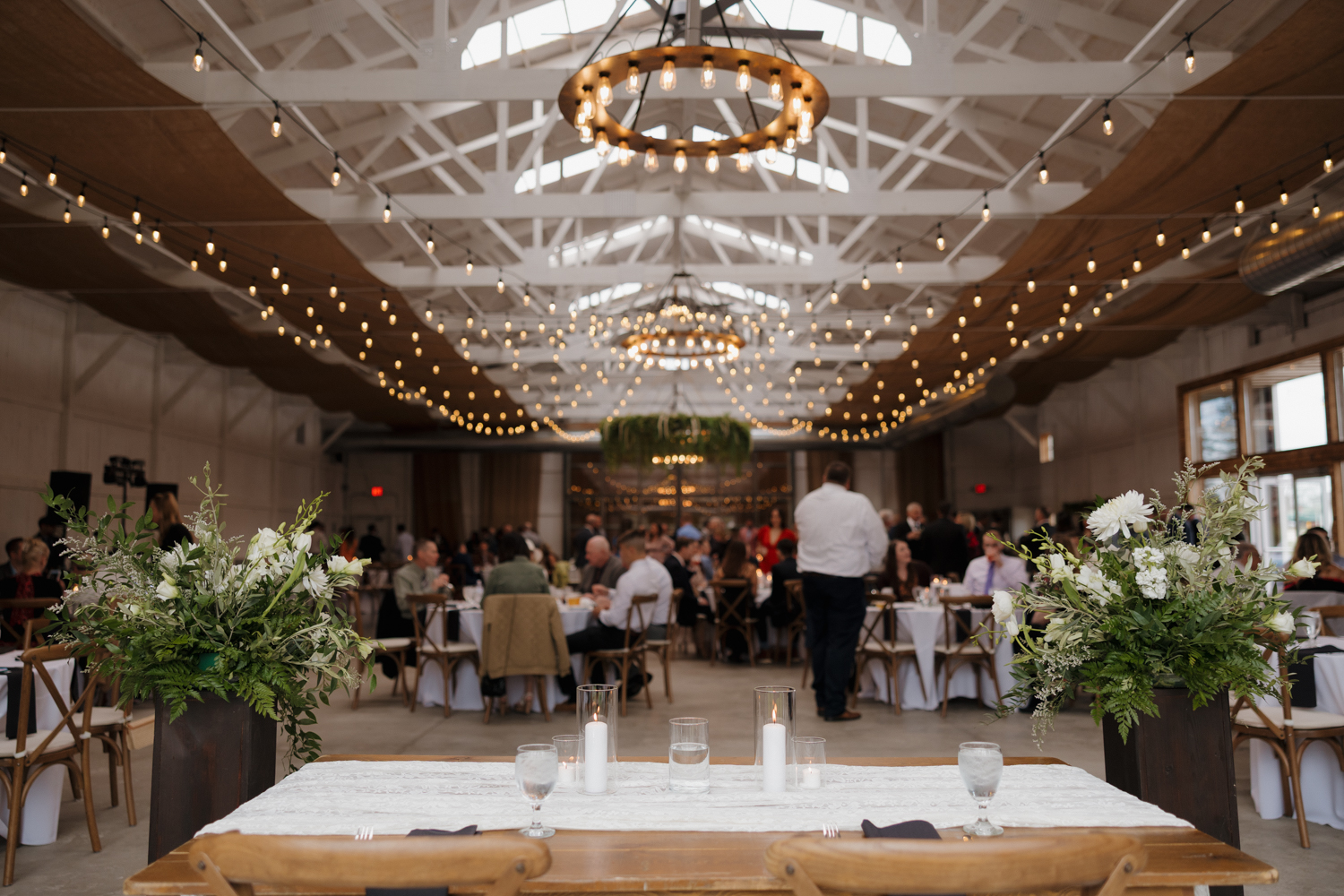 Wide view of the reception inside Boxed and Burlap Delavan with string lights draped across white beams and guests seated at round tables.