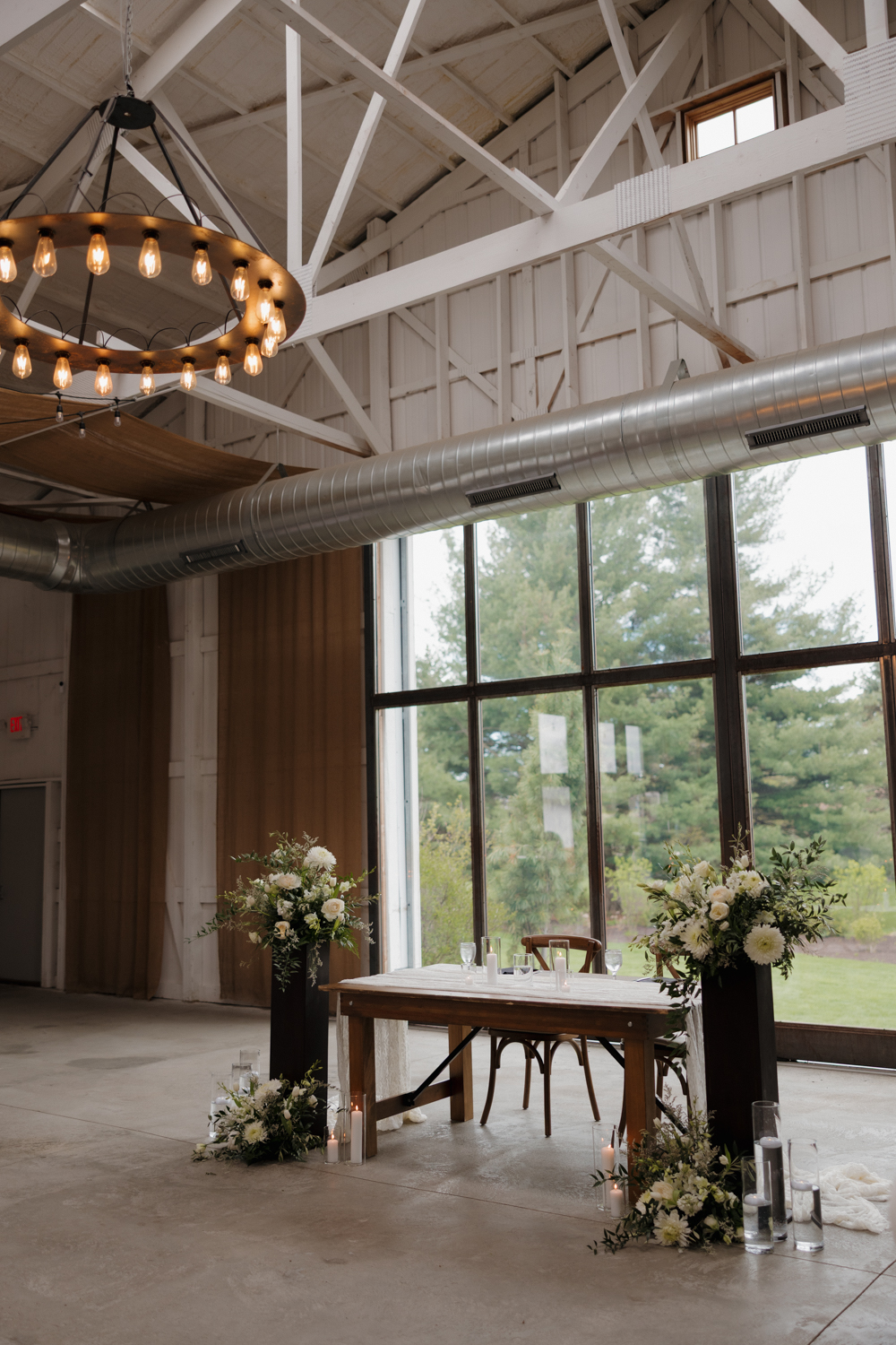 Indoor ceremony space at Boxed and Burlap Delavan featuring floor-to-ceiling windows, exposed white beams, and modern chandeliers.
