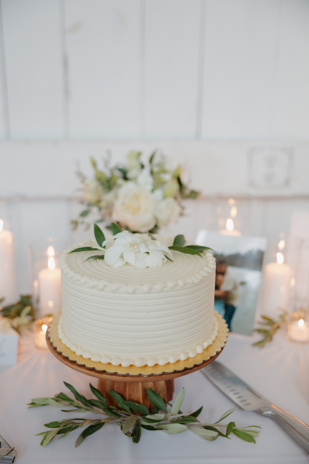 Simple white buttercream wedding cake with fresh greenery accents displayed on a wooden stand.