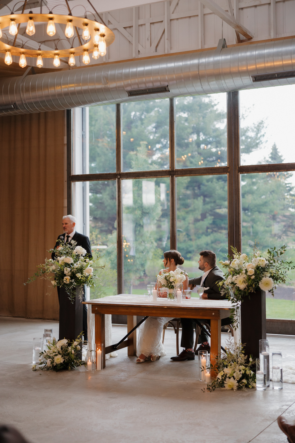 Reception toasts inside Boxed and Burlap Delavan with floor-to-ceiling windows, white beams, and lush floral arrangements framing the head table.