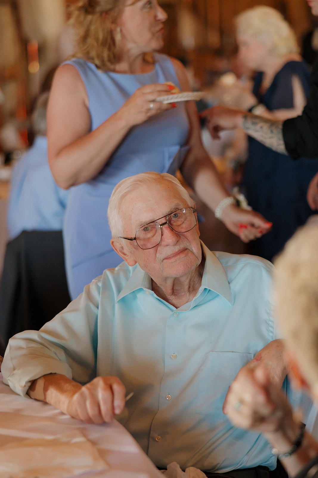 An elderly guest sits at a reception table smiling thoughtfully while wedding guests mingle in the background during a heartfelt celebration.