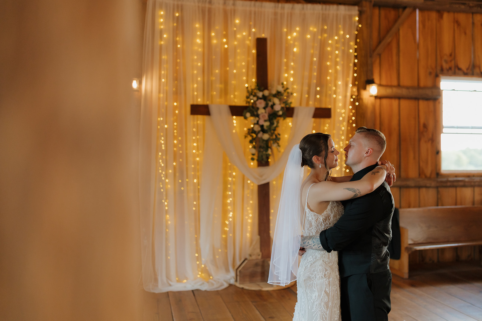 The bride and groom embrace and smile at each other beneath soft drapery and string lights inside a rustic barn, captured by a madison wisconsin wedding photographer.